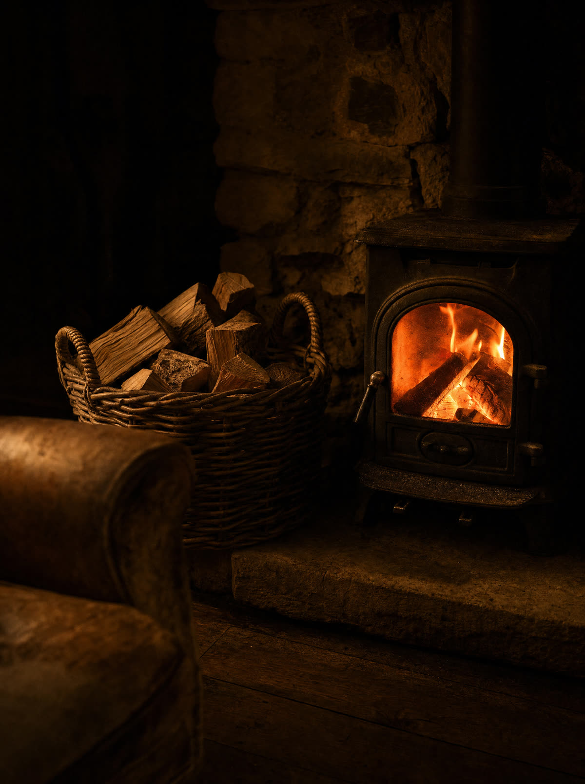 A wicker basket of split logs beside a small lit wood burner in a sitting room